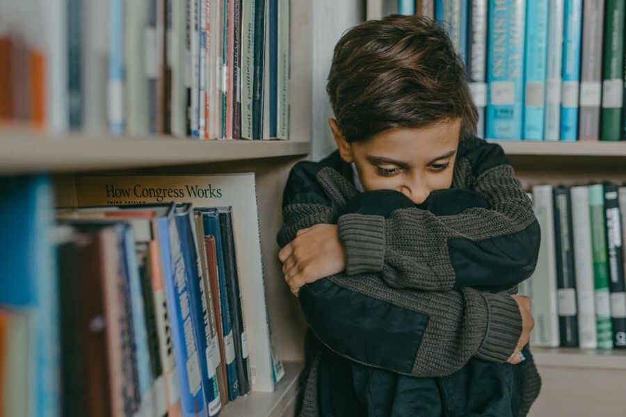A young boy sits curled up on the floor of a library, hugging his knees and looking down with a sad expression.