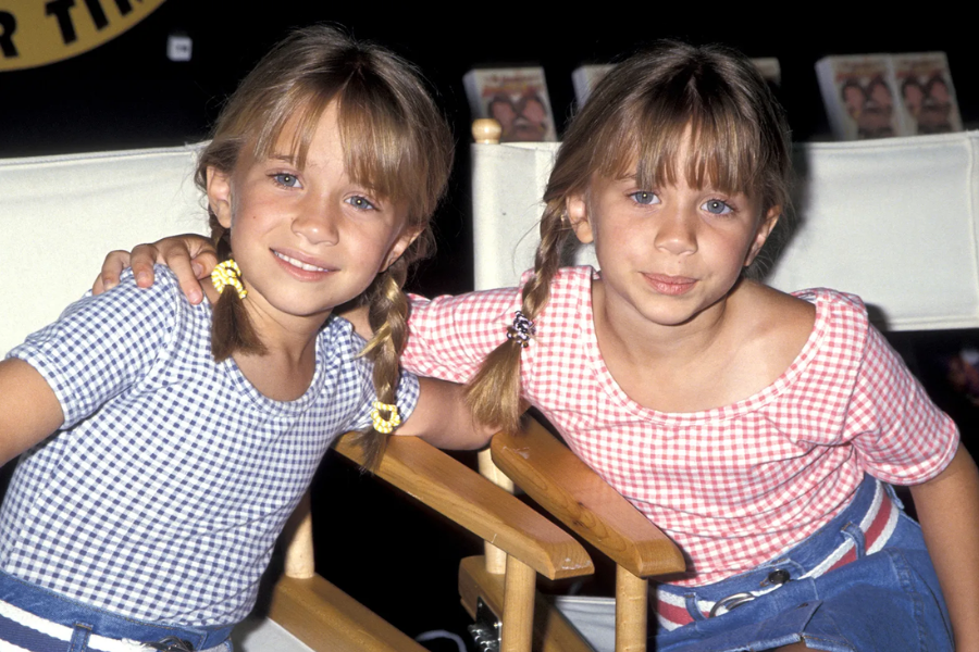 Two young girls with braided pigtails pose together in matching denim skirts and gingham tops, smiling while seated on director chairs.