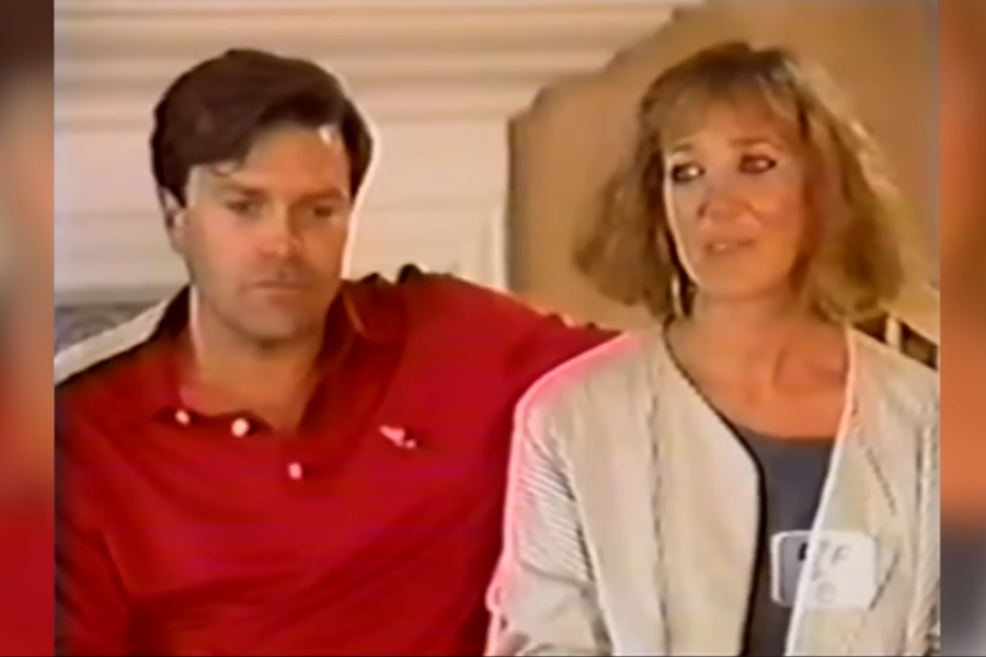 A man in a red polo shirt sits with his arm around a woman during a formal interview, both looking serious. The lighting and setup suggest a documentary or television special.