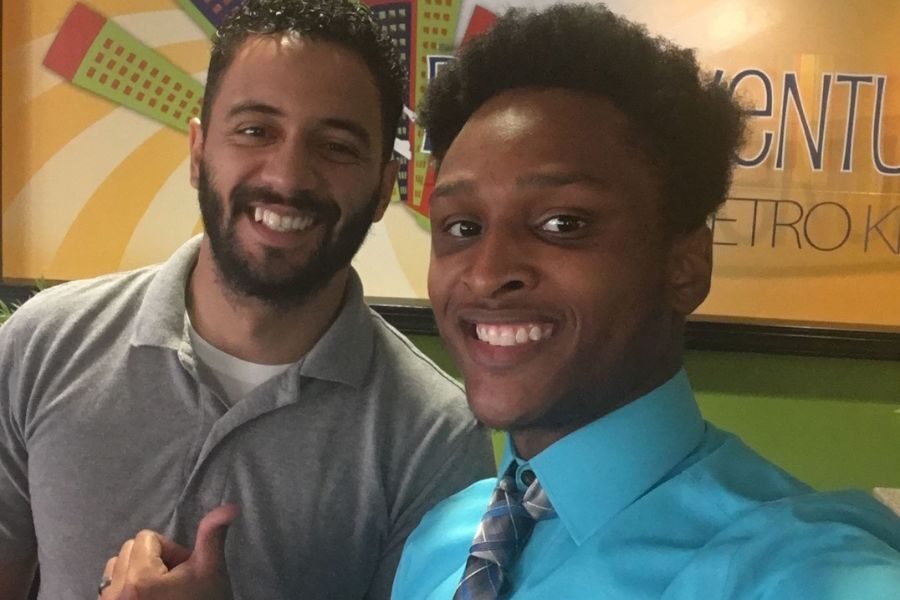 wo smiling men take a selfie indoors, one in a grey polo shirt and the other in a blue shirt and patterned tie, with colorful wall art in the background.