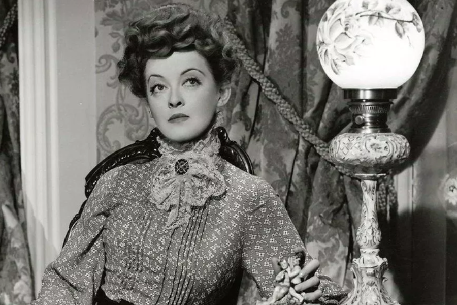 A woman in a high-collared dress sits beside a floral lamp, holding a porcelain figurine and gazing upward in a black-and-white period scene.