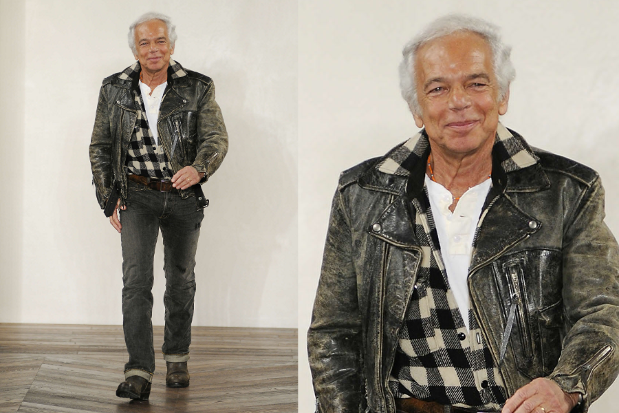 Man walking confidently in a distressed leather jacket layered over a black-and-white buffalo plaid shirt and dark jeans, on a wooden floor runway. Close-up shows his silver hair and relaxed smile.