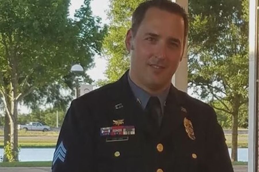 A man in a formal police uniform stands outside with trees in the background, looking at the camera with a slight smile.