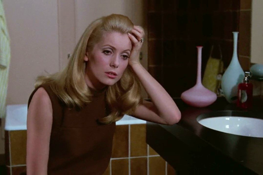 A blonde woman in a sleeveless brown dress leans on a bathroom counter with a distant, weary expression, surrounded by colorful glass vases and a sink.