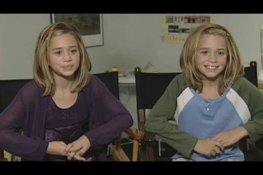 Preteen Mary-Kate and Ashley Olsen sit side by side in a casual interview setting, one wearing purple and the other in a blue and green raglan shirt, both smiling.