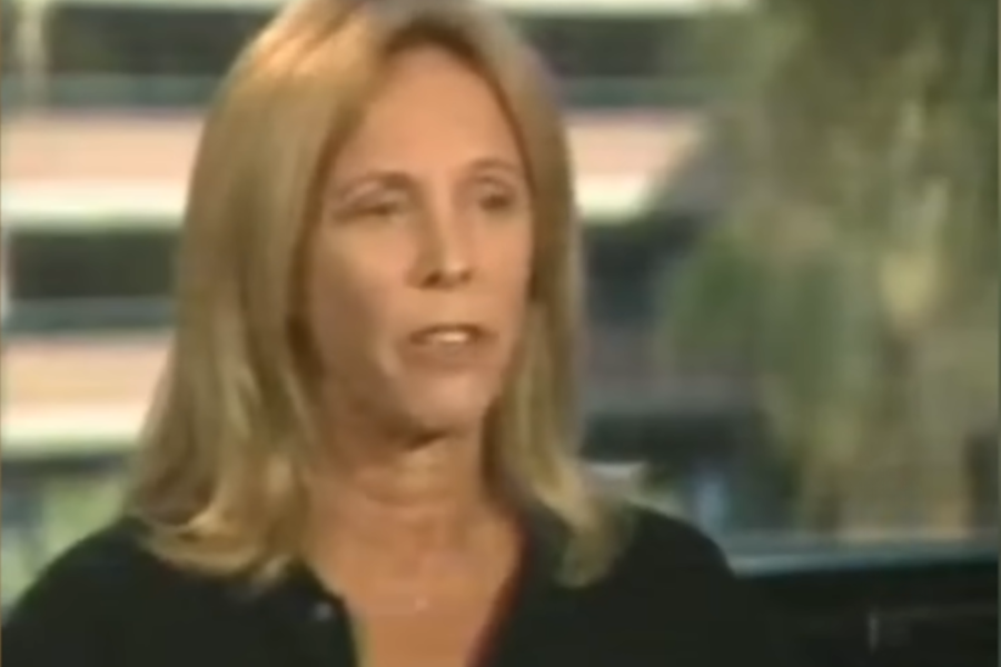 A blonde woman with straight hair and a neutral expression speaks during an interview, set against a blurred office or urban background.