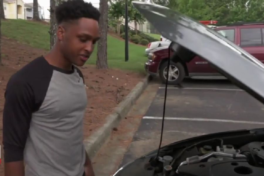 A man in a grey and black shirt looking under the open hood of a parked car in a residential area.