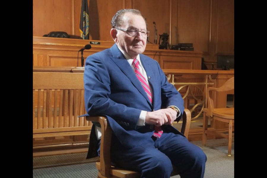 Judge Frank Caprio sits in a well-lit courtroom, dressed in a navy blue suit and red-striped tie, posing formally for what appears to be an interview or profile piece.
