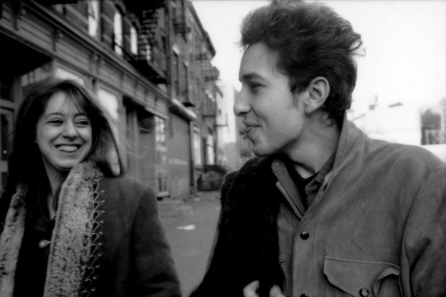 A candid black and white street photo of a smiling woman and a man mid-conversation, walking down a city sidewalk.