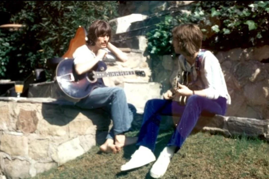 George Harrison and Eric Clapton sit barefoot on stone steps in a sunlit garden, each holding a guitar during a casual jam session.
