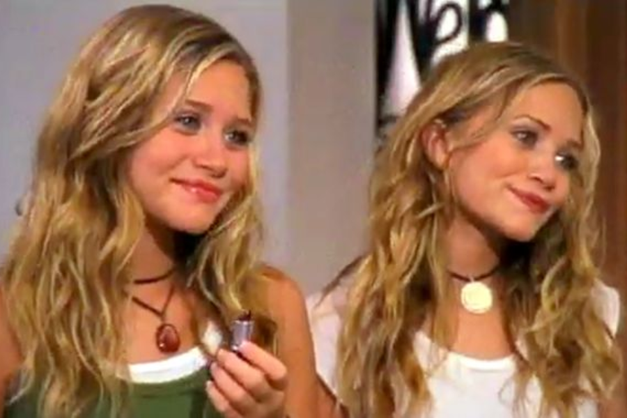 Teenage Olsen twins with long, wavy blonde hair wear trendy necklaces and casual tops, smiling in a warmly lit room.