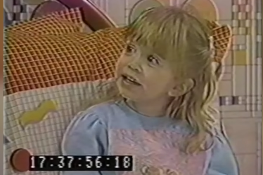 A young blonde girl with bangs sits in a colorful bedroom, wearing a pastel blue sweater with a pink patch on the front. She looks up while speaking, captured mid-sentence.
