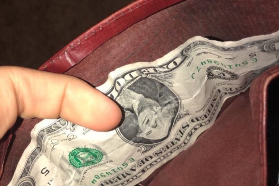 A close-up of a hand holding an open wallet containing a single crumpled one-dollar bill.