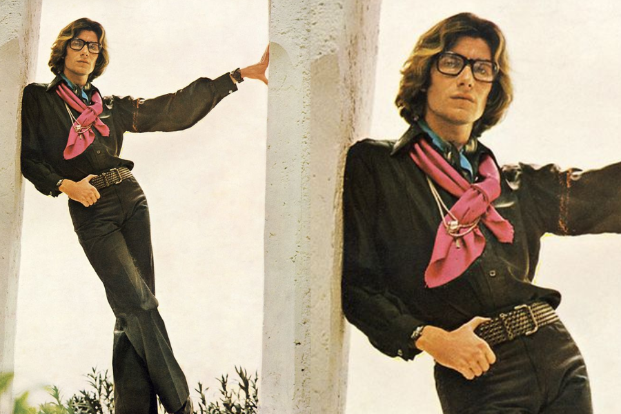 Stylish man wearing a black satin shirt and high-waisted trousers leans against a column, with layered silk scarves tied at the neck. Close-up reveals large glasses and tousled hair.