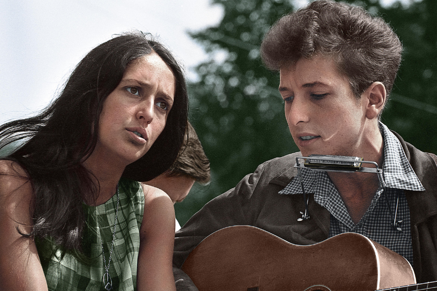 Colorized image of Joan Baez and Bob Dylan performing outdoors, Baez looking intently ahead while Dylan plays guitar and harmonica.