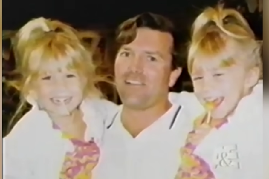 A man smiles while holding two giggling twin girls in ponytails. The girls are wearing colorful outfits and laughing together.