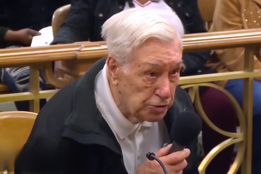 An elderly man with white hair and a serious expression speaks into a handheld microphone while seated in a courtroom gallery.