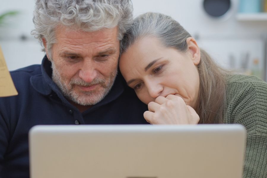 A concerned older couple leans in closely, staring at a laptop screen while holding documents, suggesting stress or worry.