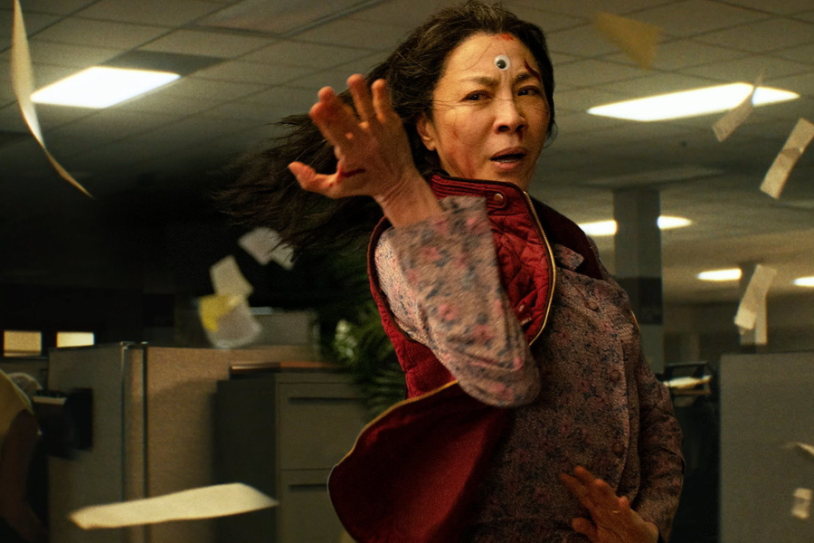 A woman in a patterned blouse and red vest strikes a martial arts pose mid-motion, surrounded by flying papers in an office, with a googly eye stuck to her forehead.