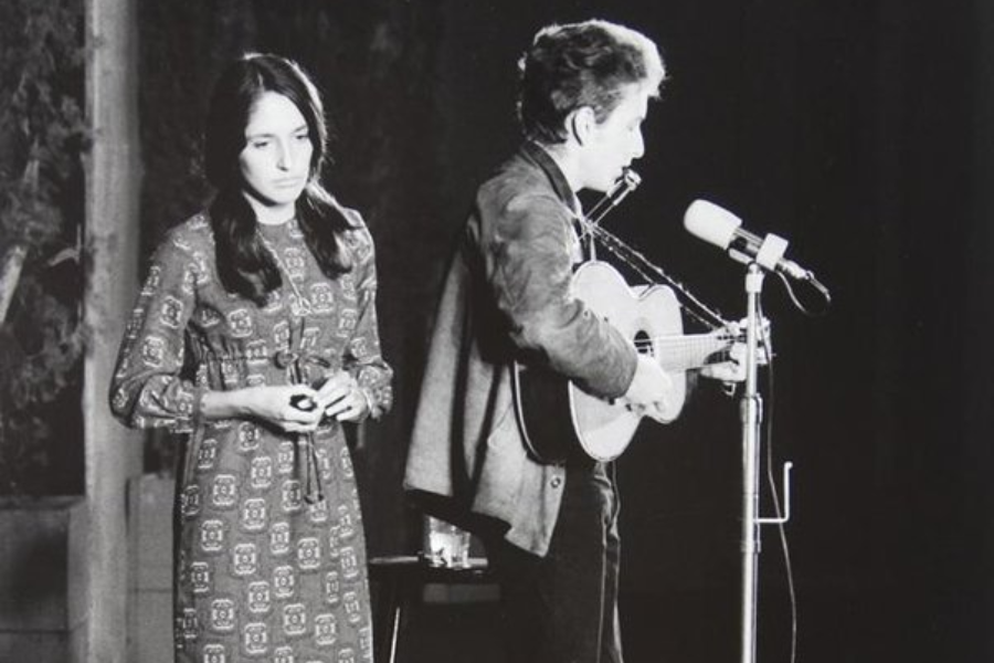 Black and white image of Joan Baez standing solemnly next to Bob Dylan, who is performing at a microphone with a guitar and harmonica.