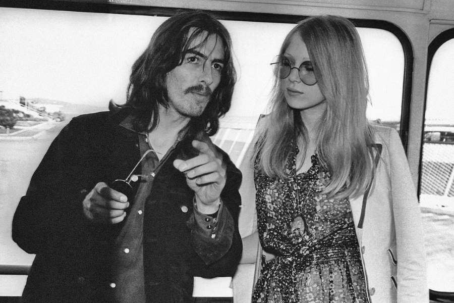 George Harrison points toward the camera while standing next to a sunglasses-wearing Pattie Boyd aboard a bus or train, both dressed in 70s fashion.