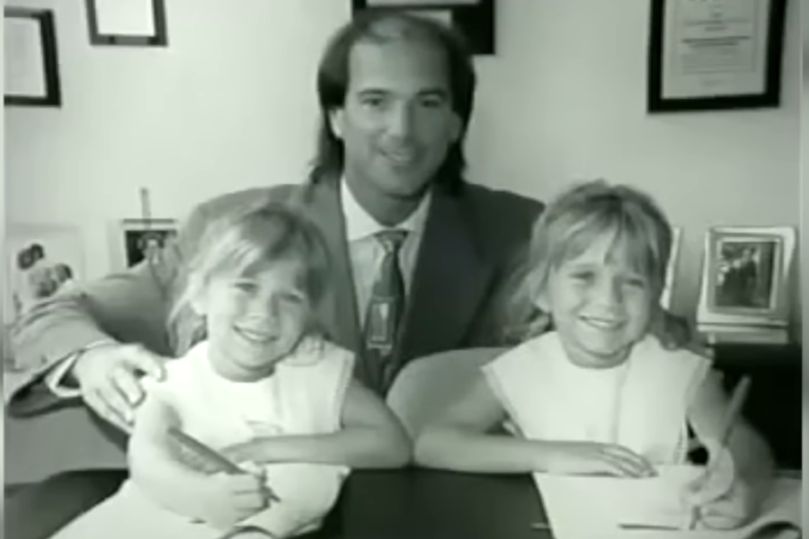 A suited man sits with two twin girls who are smiling as they sign papers at a desk. The black-and-white photo suggests a business moment or legal event.
