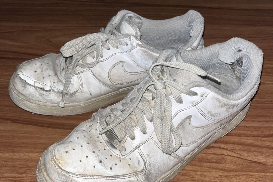 A pair of white Nike sneakers with heavy scuff marks, creases, and frayed edges, resting on a wooden floor.