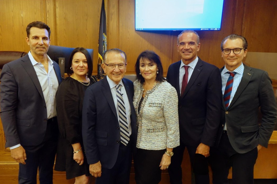 Judge Frank Caprio stands smiling in a courtroom surrounded by five adults dressed in business attire, all posing together in front of the judge’s bench with a large screen and wood-paneled wall behind them.