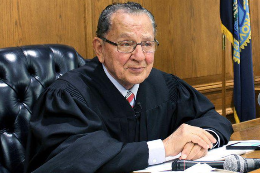 Judge Frank Caprio sits at the bench in his courtroom, hands folded with a calm and thoughtful expression, dressed in his black judicial robe. A microphone and paperwork lie in front of him, with the American and Rhode Island state flags visible in the background.