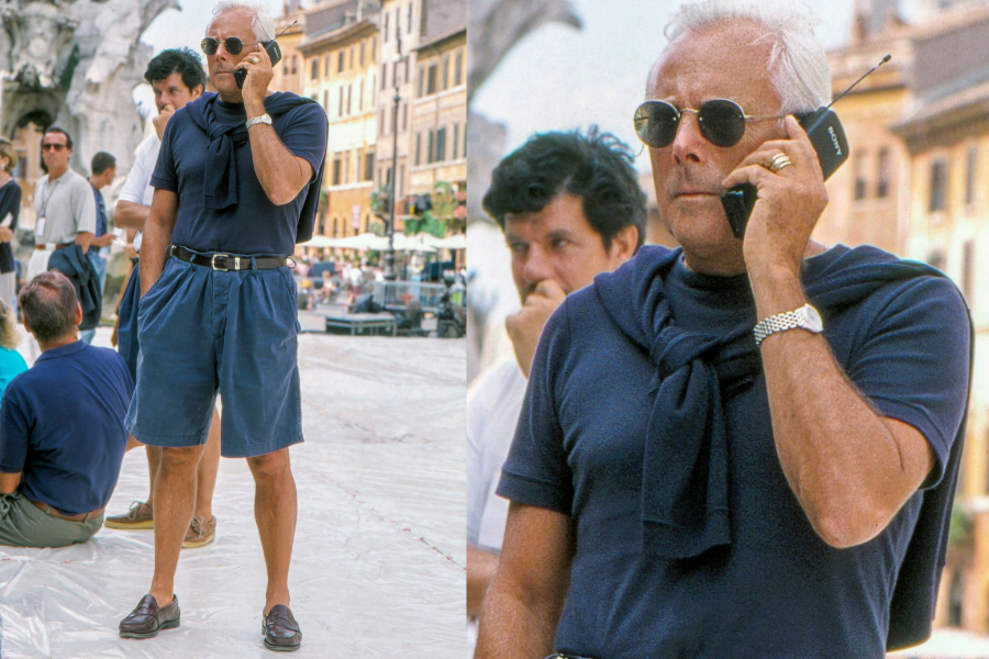 Older man in a monochrome navy outfit with belted shorts, loafers, and a sweater tied around his shoulders, speaking on a chunky 90s-style mobile phone. He wears round sunglasses and a silver watch, standing confidently in a European plaza with people in the background.