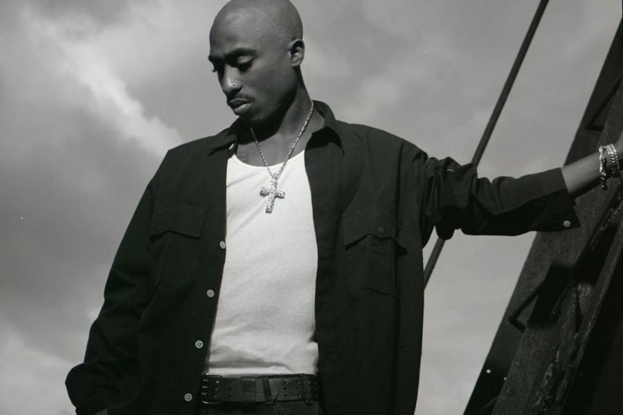 A man in a sleeveless shirt and open jacket stands pensively against a rail with a cross necklace. The sky is cloudy, creating a reflective mood.
