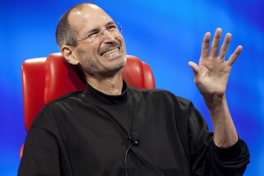 A person wearing glasses and a black turtleneck smiles warmly, seated on a red chair against a blue background, waving their hand in a friendly gesture.