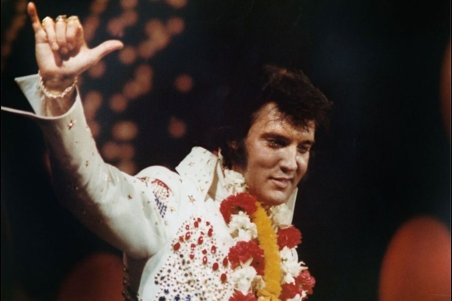 An entertainer in a white, jeweled jumpsuit and floral lei smiles warmly, raising a hand in a lively pose against a dark, blurred background.