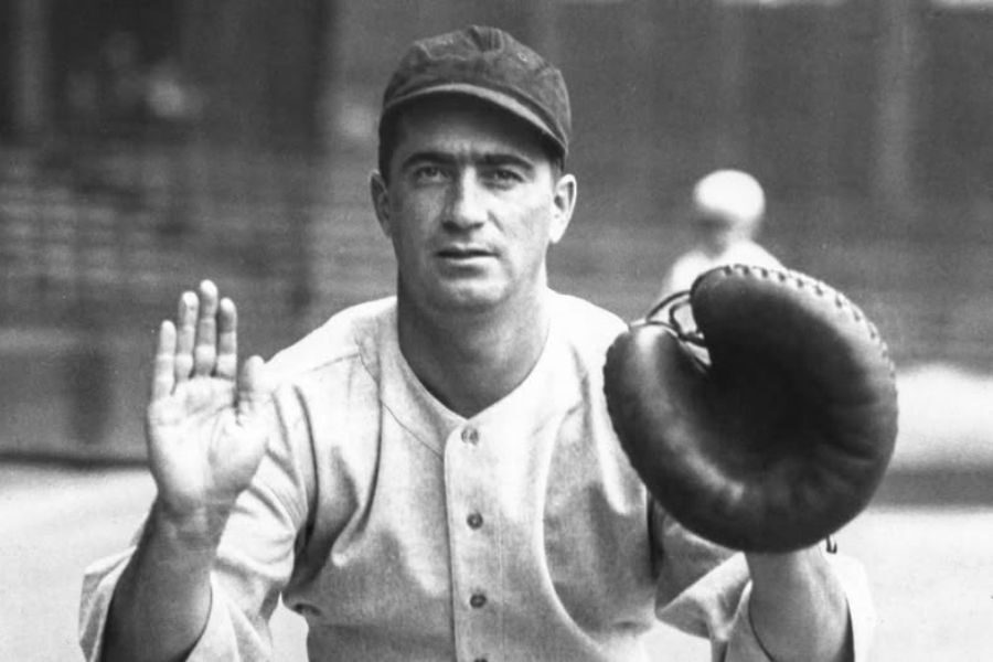 A vintage photo of a baseball player, wearing an old-fashioned uniform and cap, holding a catcher's mitt and raised hand, set in a blurred field background.