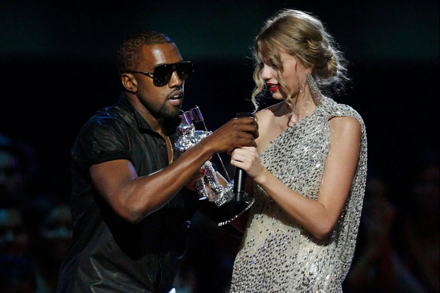 Man in sunglasses interrupting a woman in a sparkling dress on stage, holding a microphone and trophy. The atmosphere is tense and unexpected.