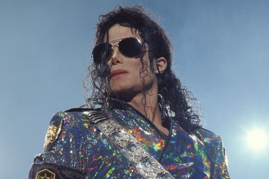 A performer with curly hair and sunglasses wears a shimmering, sequined jacket and holds a microphone on stage, exuding a confident presence.