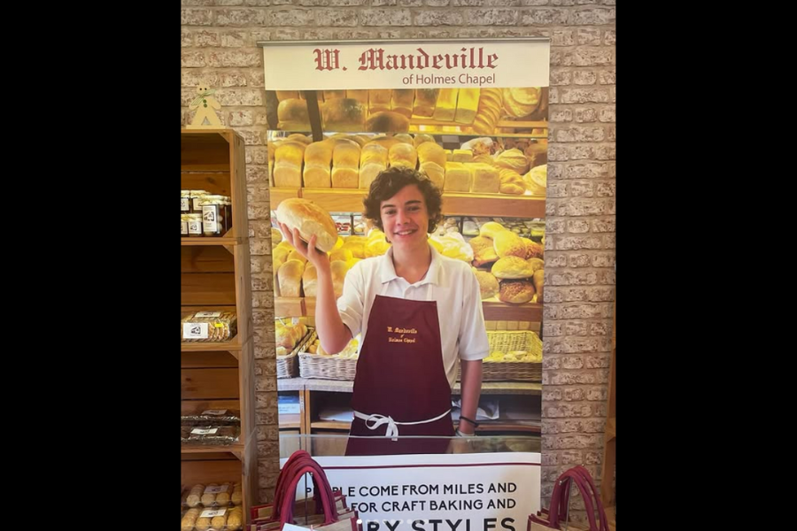 A bright, welcoming bakery display featuring a young person in a maroon apron, holding a loaf of bread. Shelves behind are lined with numerous golden-brown loaves, creating a warm, inviting atmosphere.