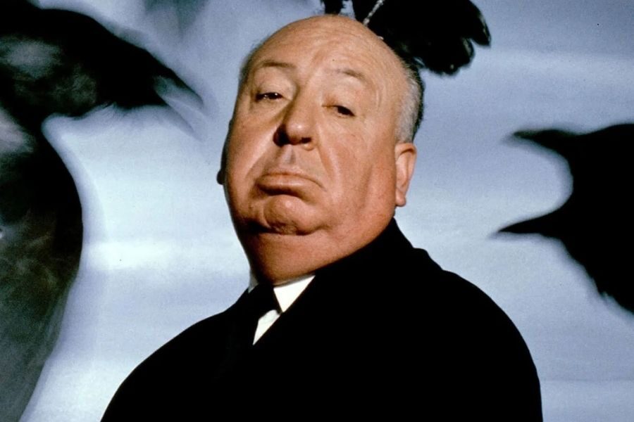 A man in a suit with a calm expression stands against a sky backdrop. Birds are blurred in motion around him, conveying suspense and intrigue.