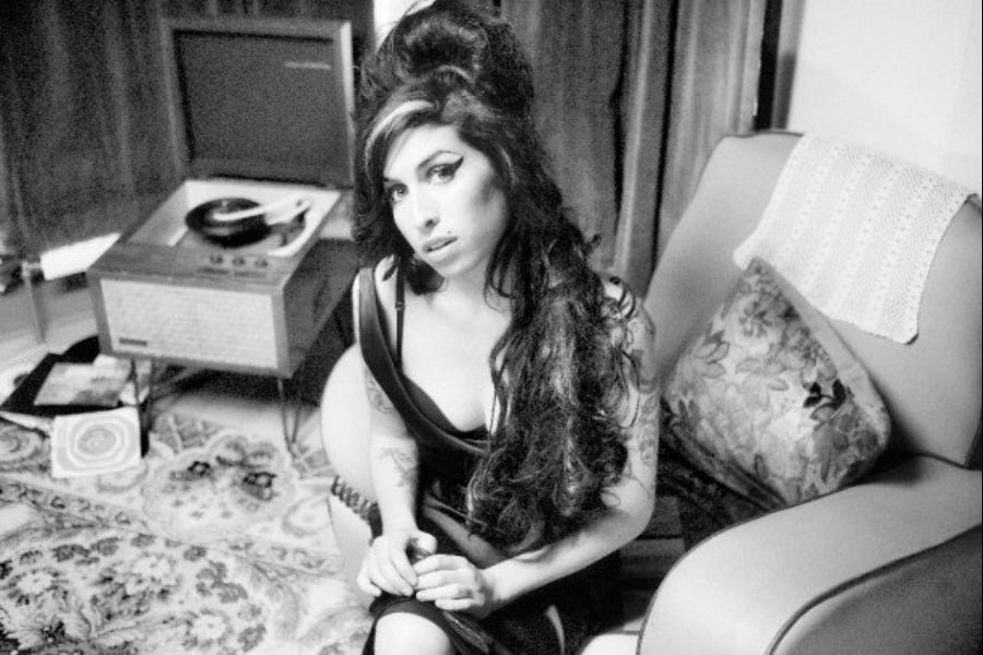 A woman with long, dark hair styled in a beehive sits thoughtfully in a vintage-inspired room. An old record player is in the background, evoking a nostalgic mood.