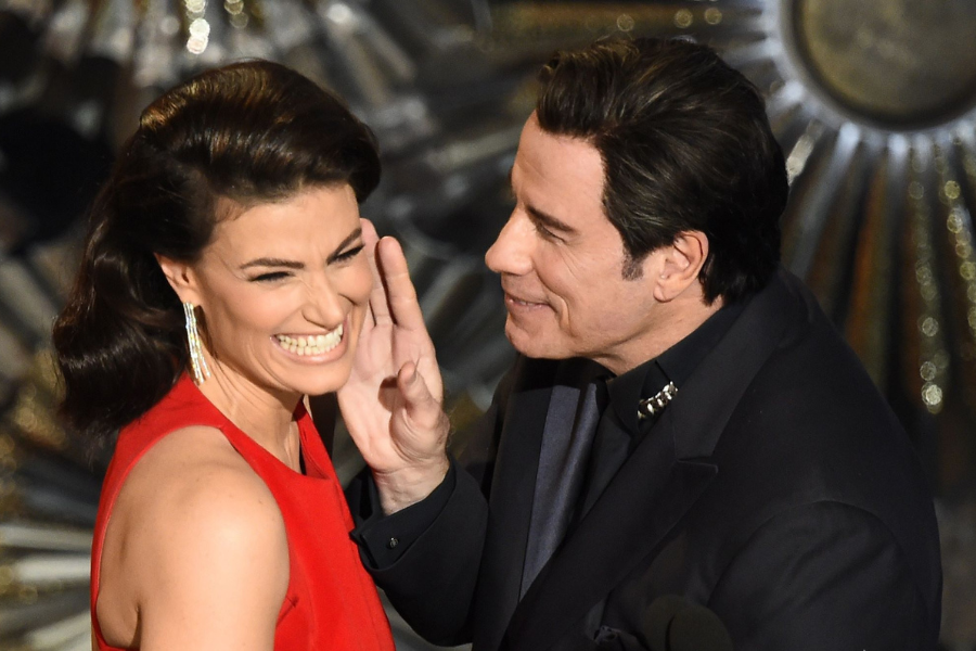 A woman in a red dress smiles broadly while a man in a dark suit gently holds her chin on a stage. The atmosphere appears lighthearted and playful.