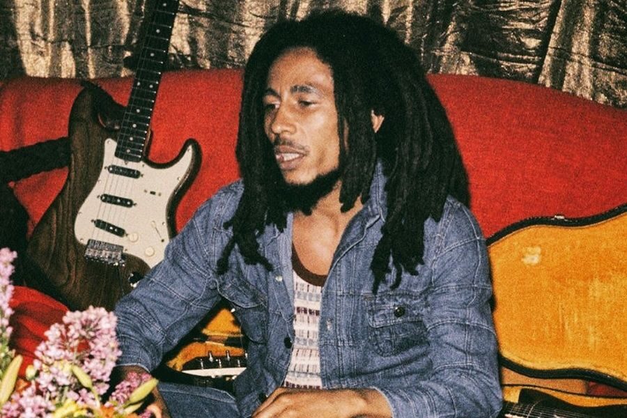 Man with long dreadlocks, wearing a denim shirt, sits on a red couch beside a guitar. A relaxed, artistic atmosphere with flowers in the foreground.