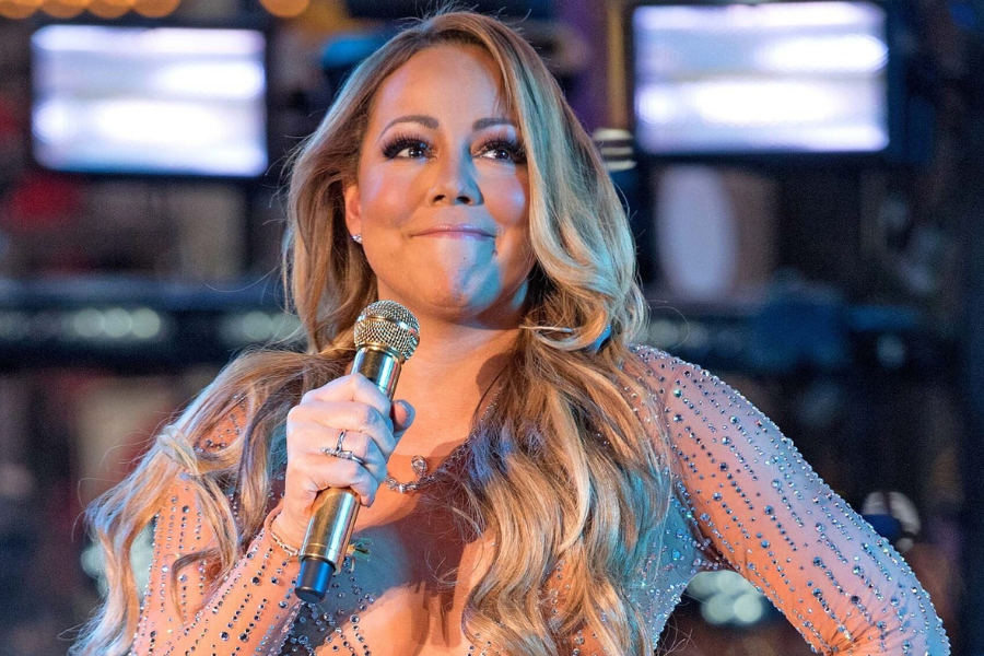 A person with long, wavy hair holds a microphone on stage, wearing a sparkly outfit. They appear thoughtful, with bright lights in the blurred background.