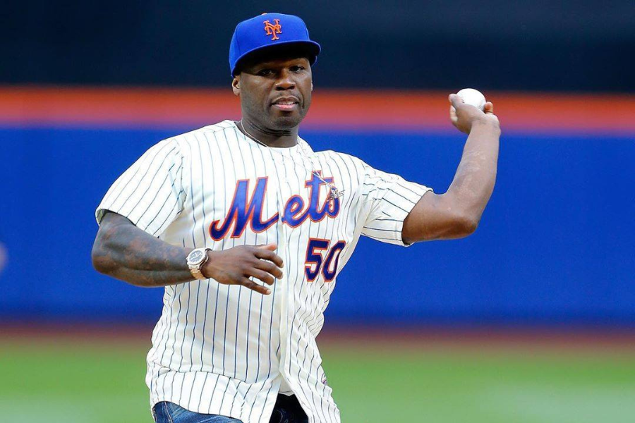 Man in a Mets jersey and cap throws a baseball on a field. He appears focused, with a crowd and a vibrant stadium background visible.