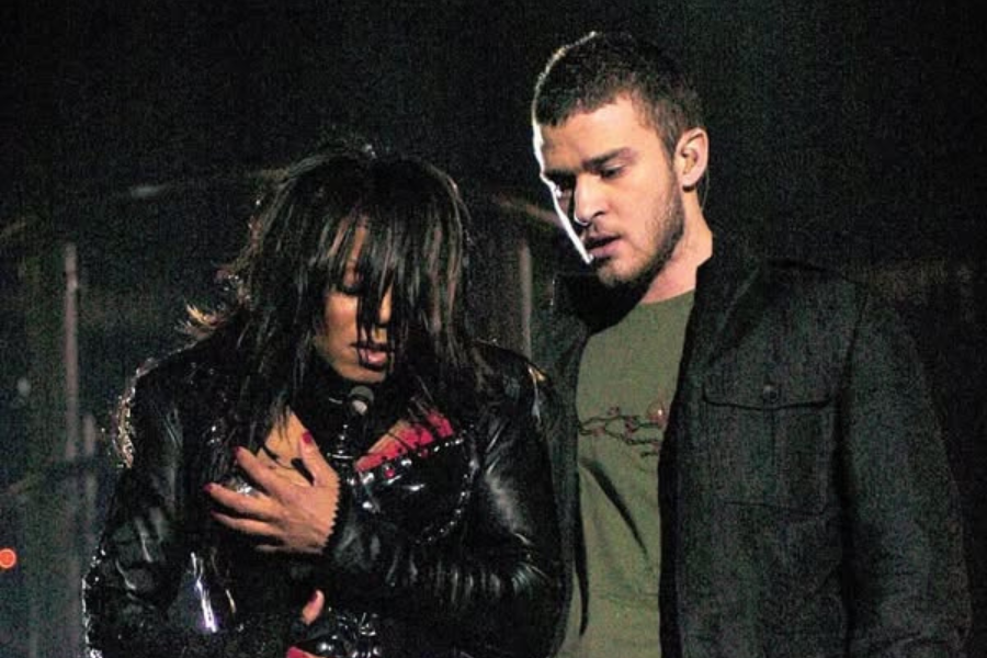 A man and woman stand on stage under dim lights. The woman holds her chest, looking down with long hair obscuring her face. The man gazes towards her, appearing concerned.