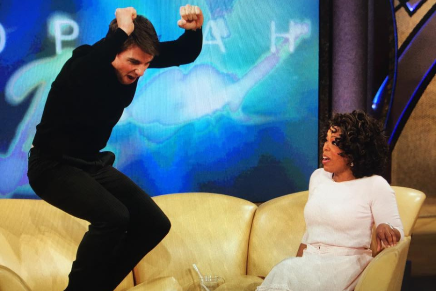 A man in black joyfully jumps on a yellow couch next to a seated woman in white. She appears surprised. A colorful, abstract backdrop is behind them.