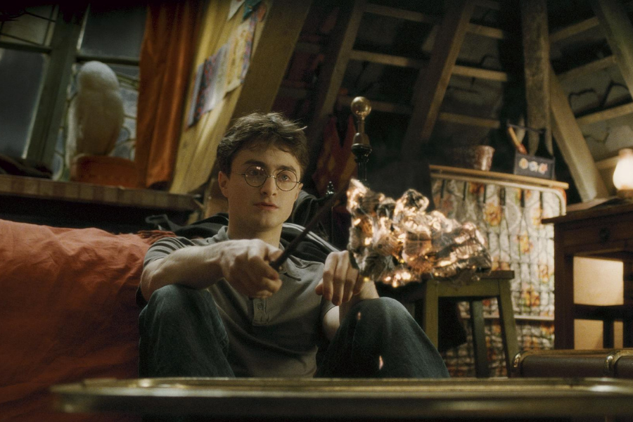 A young man with glasses sits on a couch holding a wand, casting a spell with sparks flying. A cozy room with an owl in the background.