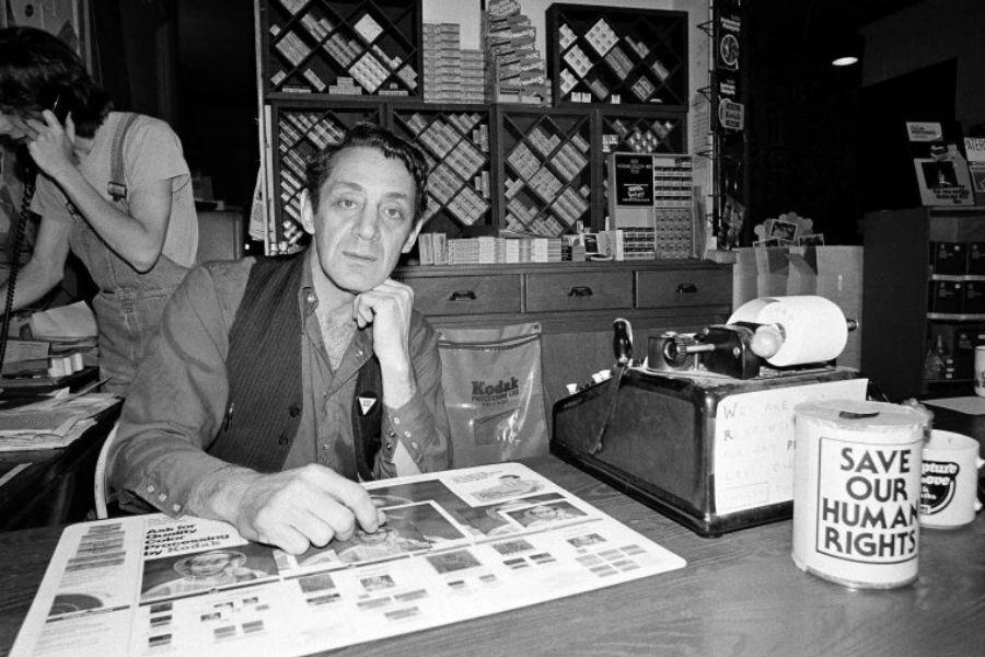 Black and white image of a man seated at a desk, hand on chin, with a cash register and "Save Our Human Rights" can. Background shows shelves with film.