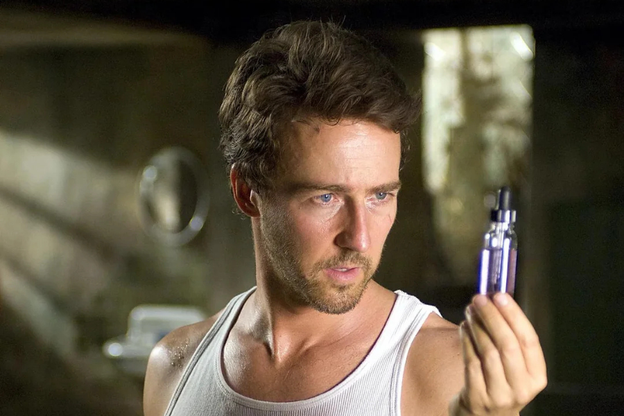 A man in a dim room intensely examines a small purple vial. He wears a white tank top, and the scene is tense and contemplative, implying intrigue.