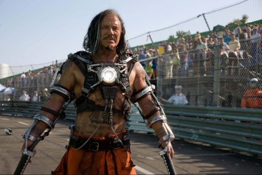 A man in rugged, metallic armor with glowing chest piece stands confidently on a racetrack. Spectators behind a fence watch the intense scene.