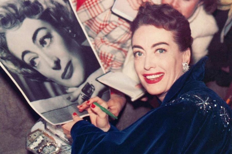 A woman in a blue velvet coat smiles brightly while signing a black-and-white photo of herself. She holds a green pen, surrounded by fans with notepads.